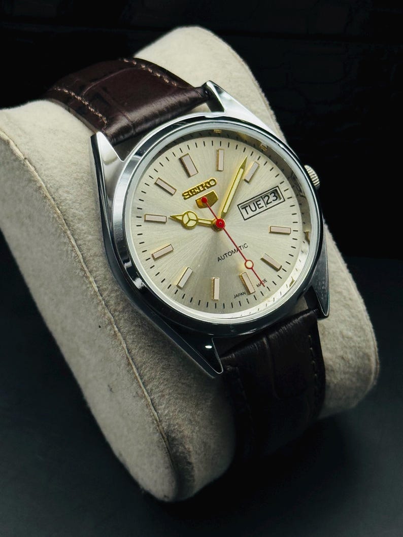 May include: Silver-toned Seiko automatic watch with a light-colored face, gold hands, and a red second hand. The watch features a day and date display and a brown leather strap. The watch is displayed on a gray cushion.