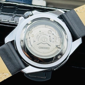 May include: A silver-toned Seiko watch with a black leather band. The watch's back is transparent, revealing the inner workings, including the text "JAPAN ENTEEN 17 JEWELS" and the Seiko logo. The watch is on a black surface.