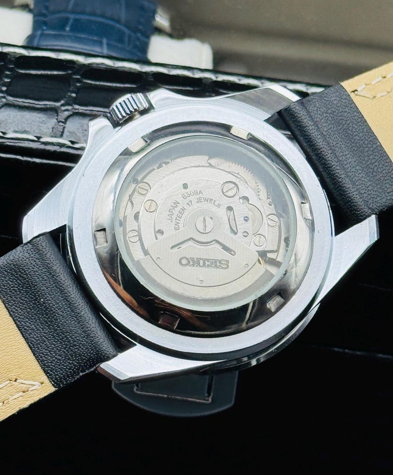 May include: A silver-toned Seiko watch with a black leather band. The watch's back is transparent, revealing the inner workings, including the text "JAPAN ENTEEN 17 JEWELS" and the Seiko logo. The watch is on a black surface.