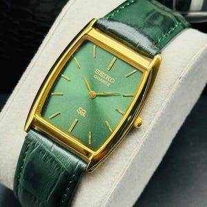 May include: A gold-toned Seiko Quartz watch with a rectangular green face and gold hands and markers. The watch has a green leather band. The watch is displayed on a white cushion.