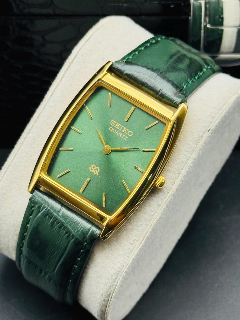 May include: A gold-toned Seiko Quartz watch with a rectangular green face and gold hands and markers. The watch has a green leather band. The watch is displayed on a white cushion.