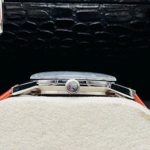 Puede incluir: Un reloj de pulsera plateado con esfera redonda y corona. El reloj tiene una esfera de color claro y una correa de cuero roja. El reloj se exhibe sobre una superficie blanca sobre un fondo oscuro.