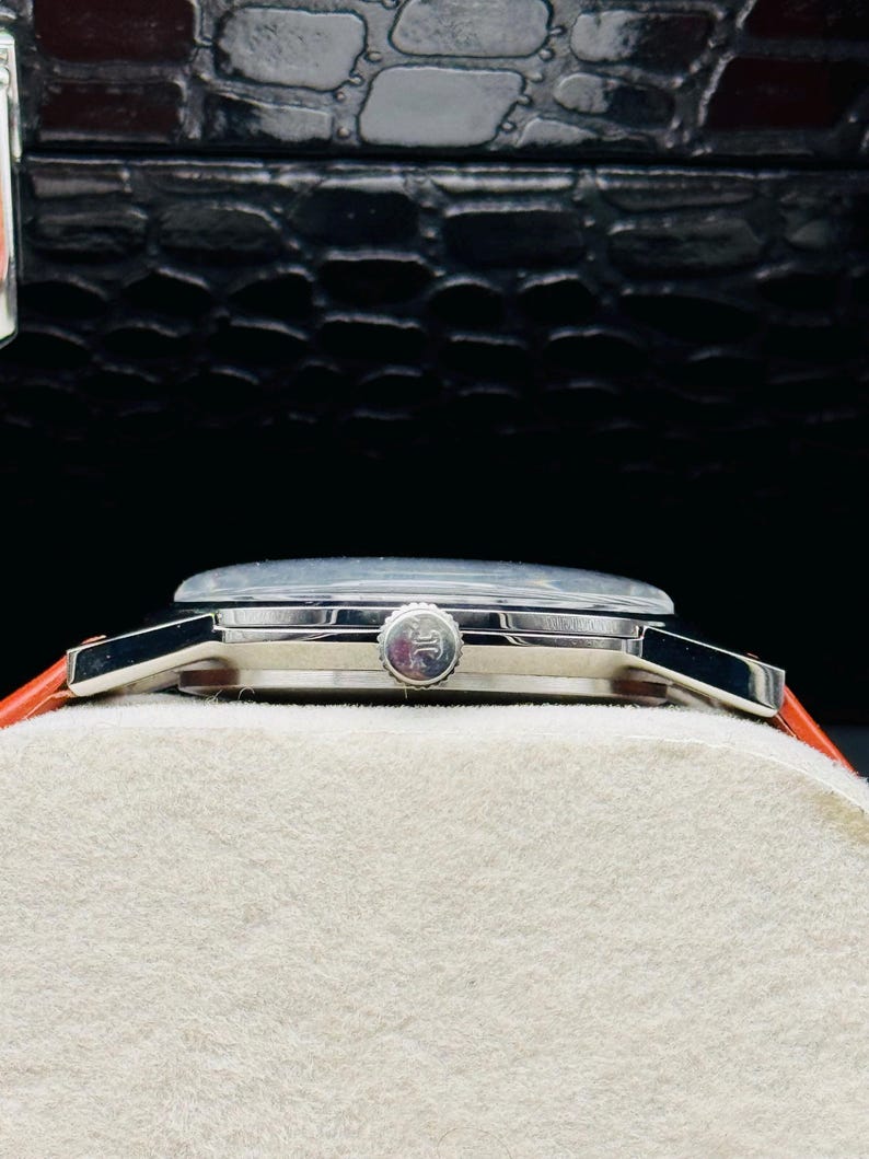 Puede incluir: Un reloj de pulsera plateado con esfera redonda y corona. El reloj tiene una esfera de color claro y una correa de cuero roja. El reloj se exhibe sobre una superficie blanca sobre un fondo oscuro.
