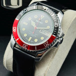 May include: A Seiko Marine Master automatic professional watch with a black dial, red bezel, and black leather strap. The watch features a date display and luminous markers. The watch is displayed on a cream-colored cushion.