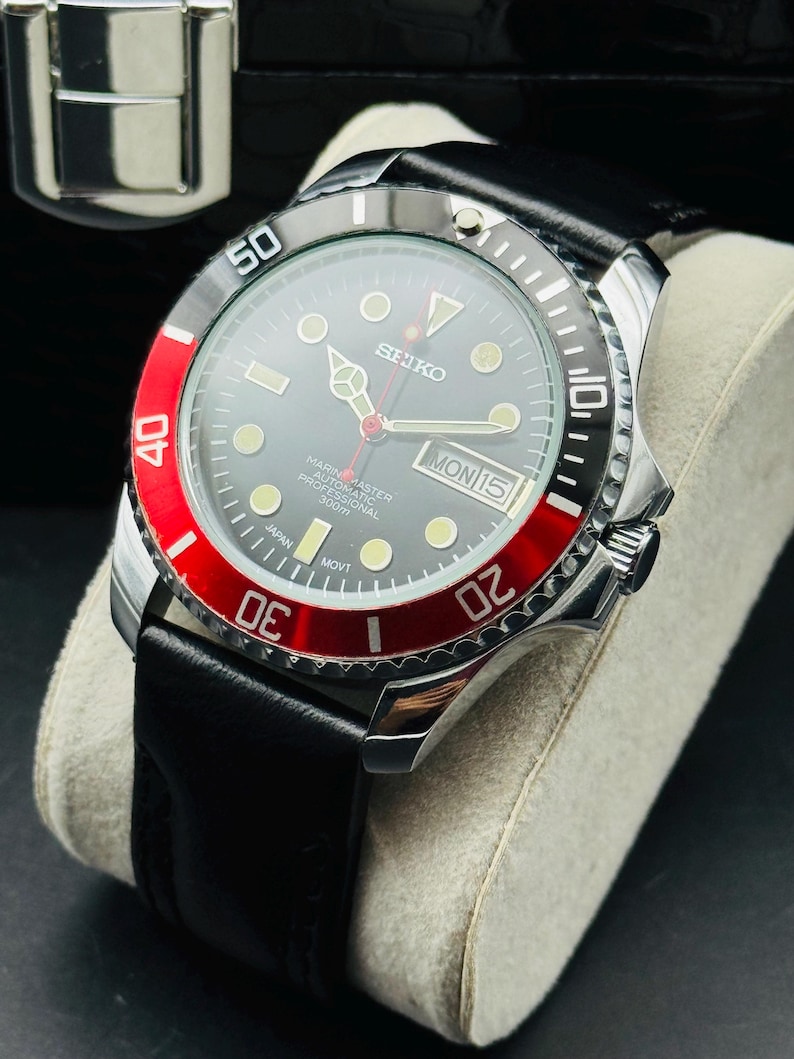 May include: A Seiko Marine Master automatic professional watch with a black dial, red bezel, and black leather strap. The watch features a date display and luminous markers. The watch is displayed on a cream-colored cushion.
