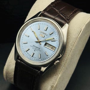 May include: A vintage Seiko 5 Sports automatic watch with a light blue dial and silver-tone hands and markers. The watch features a day and date display and a brown leather strap. The watch is displayed on a cream-colored cushion.