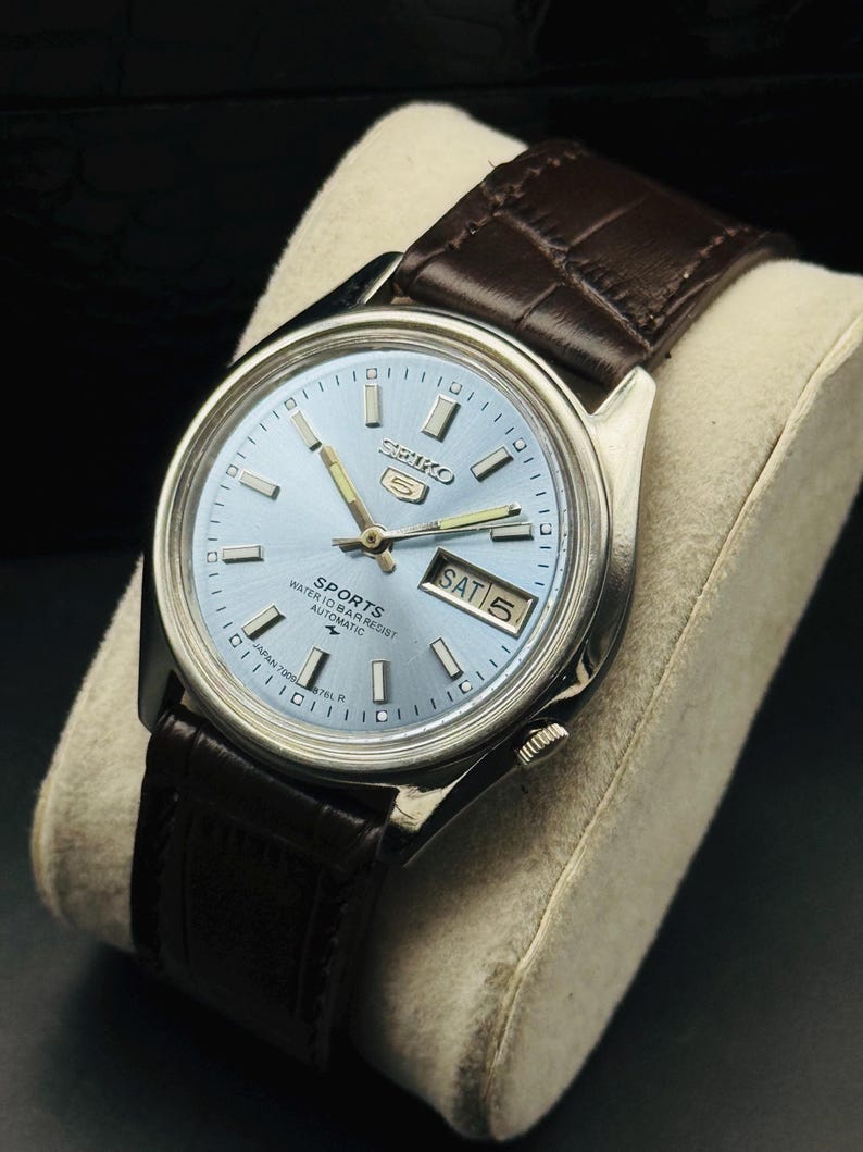 May include: A vintage Seiko 5 Sports automatic watch with a light blue dial and silver-tone hands and markers. The watch features a day and date display and a brown leather strap. The watch is displayed on a cream-colored cushion.