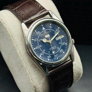 May include: A silver-cased Seiko watch with a dark blue face and a brown leather strap. The watch face displays the day and date, with white numerals and markers. The watch is an automatic with 21 jewels.
