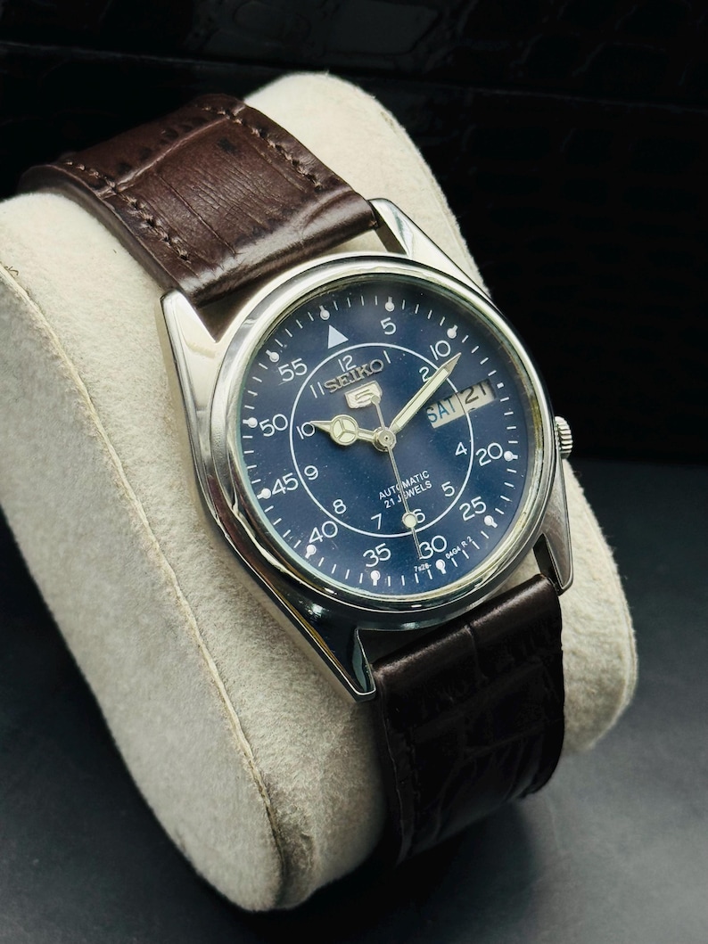 May include: A silver-cased Seiko watch with a dark blue face and a brown leather strap. The watch face displays the day and date, with white numerals and markers. The watch is an automatic with 21 jewels.