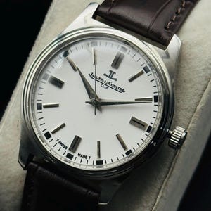 May include: A silver-toned Jaeger-LeCoultre Club watch with a white face and black markers. The watch has a brown leather band. The watch face displays the Jaeger-LeCoultre logo and the words "T Swiss Made T".