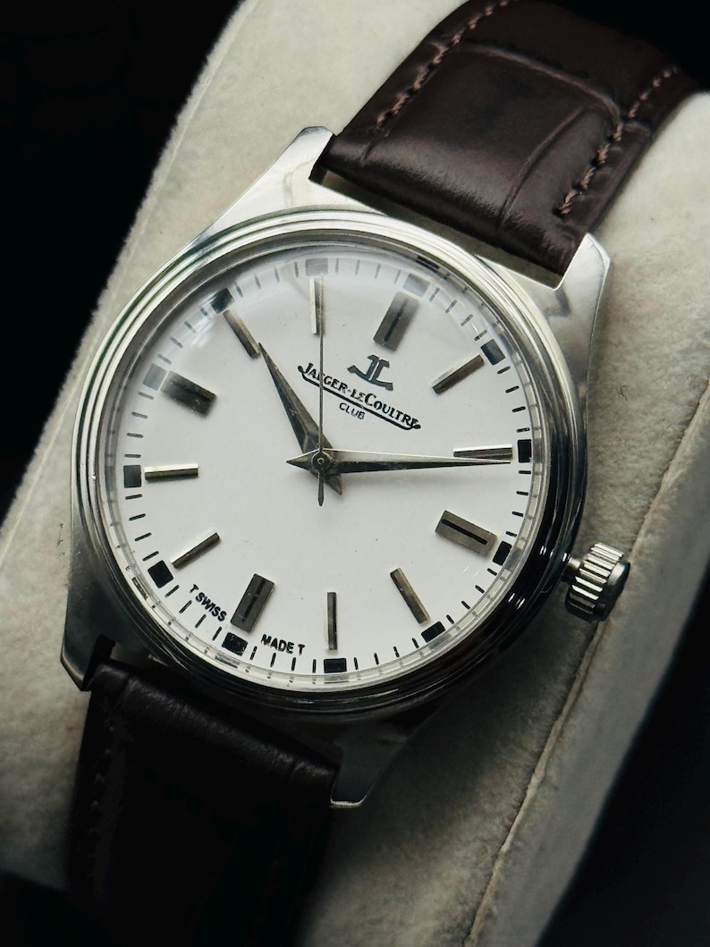 May include: A silver-toned Jaeger-LeCoultre Club watch with a white face and black markers. The watch has a brown leather band. The watch face displays the Jaeger-LeCoultre logo and the words "T Swiss Made T".