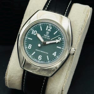 May include: A classic Tudor Oyster watch with a green face and a silver case. The watch has white hands and numerals, a black leather strap with white stitching, and the words "SHOCK RESISTING" and "SWISS 30 MADE" on the dial.