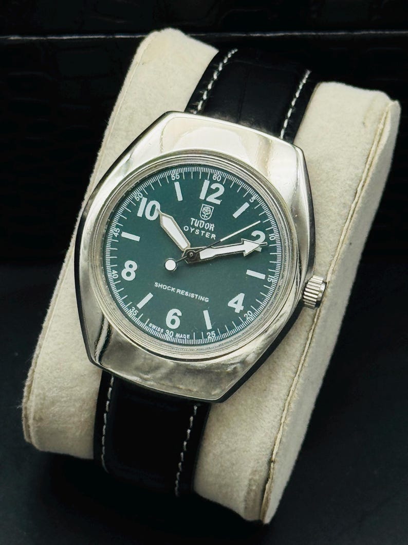 May include: A classic Tudor Oyster watch with a green face and a silver case. The watch has white hands and numerals, a black leather strap with white stitching, and the words "SHOCK RESISTING" and "SWISS 30 MADE" on the dial.