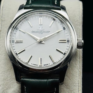 May include: A silver-toned Jaeger-LeCoultre Club watch with a white face, silver hands, and a dark green leather strap. The watch face features the Jaeger-LeCoultre logo and the words "T Swiss Made T".