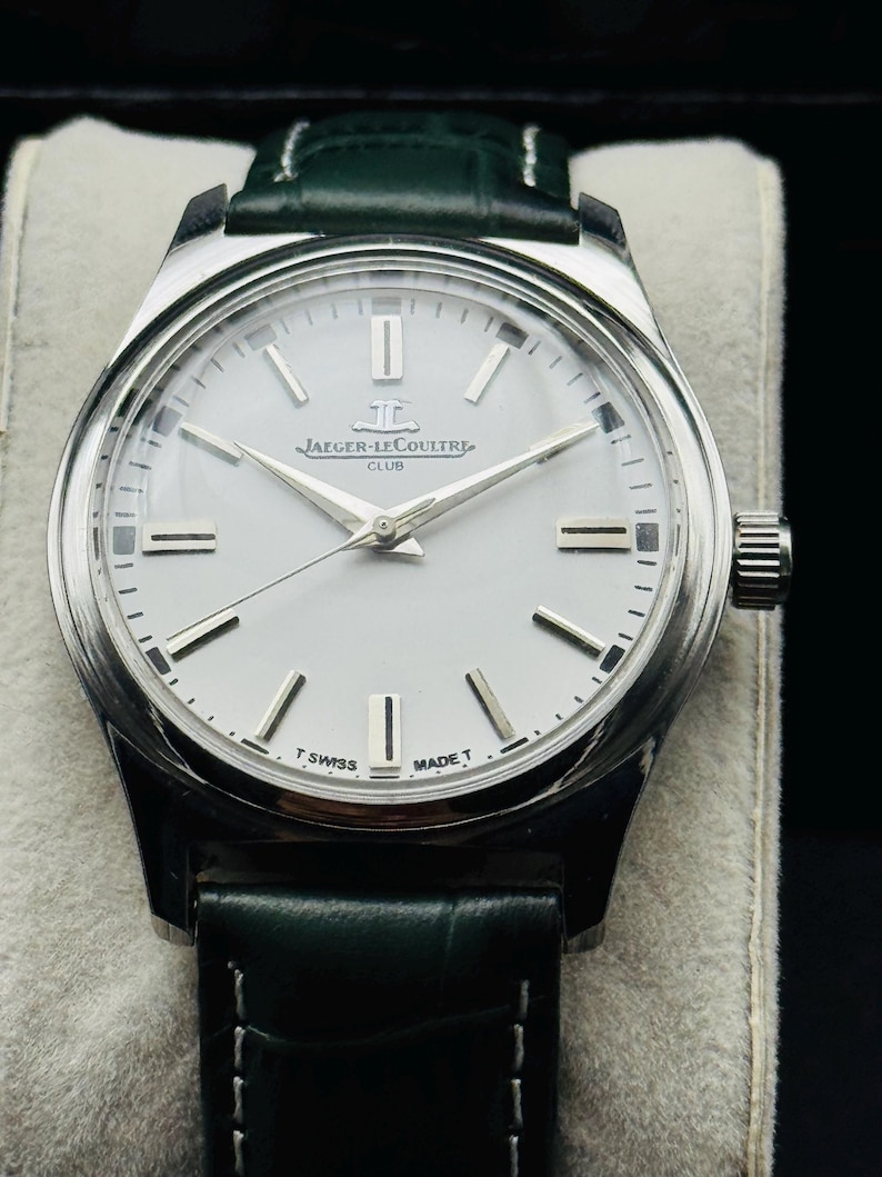 May include: A silver-toned Jaeger-LeCoultre Club watch with a white face, silver hands, and a dark green leather strap. The watch face features the Jaeger-LeCoultre logo and the words "T Swiss Made T".