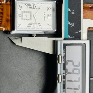 Op de afbeelding: Een rechthoekig Seiko horloge met een zilveren kast en een lichtgekleurde wijzerplaat met Romeinse cijfers. Het horloge heeft een bruine leren band. De digitale schuifmaat toont een meting van 29,77 mm.