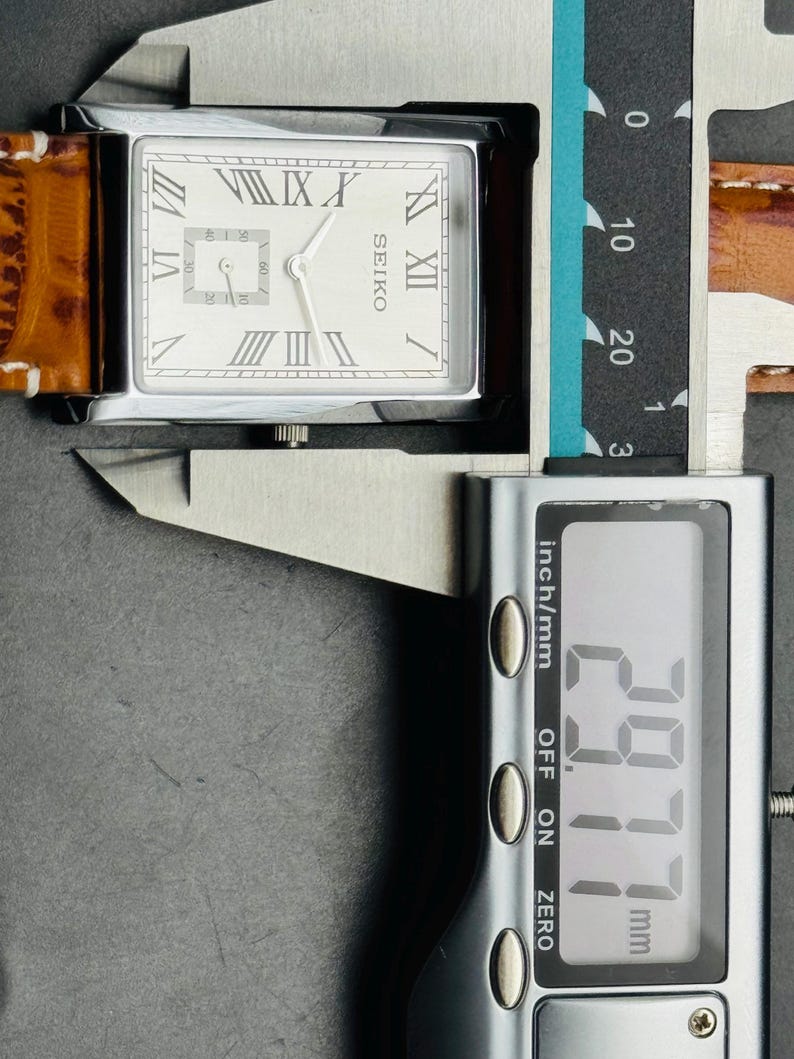 Op de afbeelding: Een rechthoekig Seiko horloge met een zilveren kast en een lichtgekleurde wijzerplaat met Romeinse cijfers. Het horloge heeft een bruine leren band. De digitale schuifmaat toont een meting van 29,77 mm.