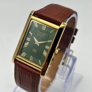 Puede incluir: Reloj Seiko Quartz rectangular con caja dorada y esfera verde. El reloj presenta n&uacute;meros romanos y agujas doradas, y una correa de cuero marr&oacute;n. El reloj se exhibe sobre un soporte transparente.