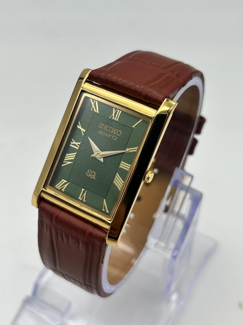 Puede incluir: Reloj Seiko Quartz rectangular con caja dorada y esfera verde. El reloj presenta n&uacute;meros romanos y agujas doradas, y una correa de cuero marr&oacute;n. El reloj se exhibe sobre un soporte transparente.