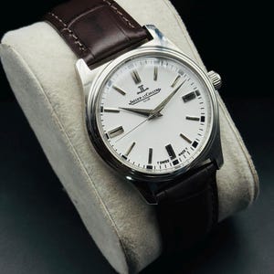 May include: A silver-toned Jaeger-LeCoultre Club watch with a white face and black markers. The watch has a brown leather band. The watch is displayed on a white cushion against a black background.