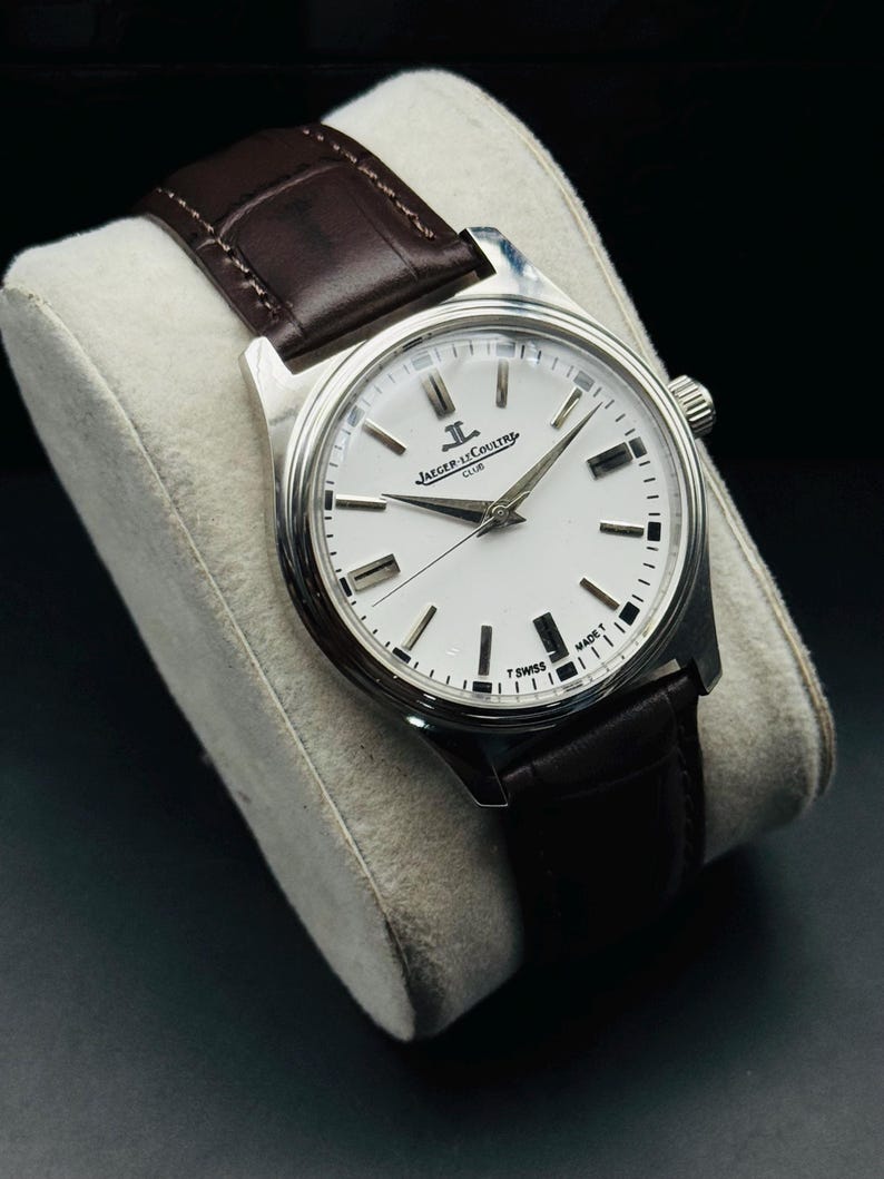 May include: A silver-toned Jaeger-LeCoultre Club watch with a white face and black markers. The watch has a brown leather band. The watch is displayed on a white cushion against a black background.