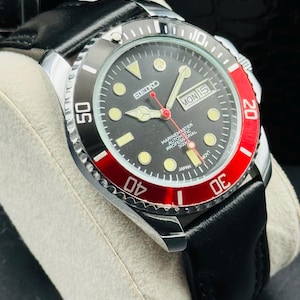 May include: A black and red Seiko Marinemaster automatic watch with a black leather strap. The watch face is black with luminous markers and hands. The bezel is black and red, with white numerals. The day and date are displayed at the 3 o'clock position.