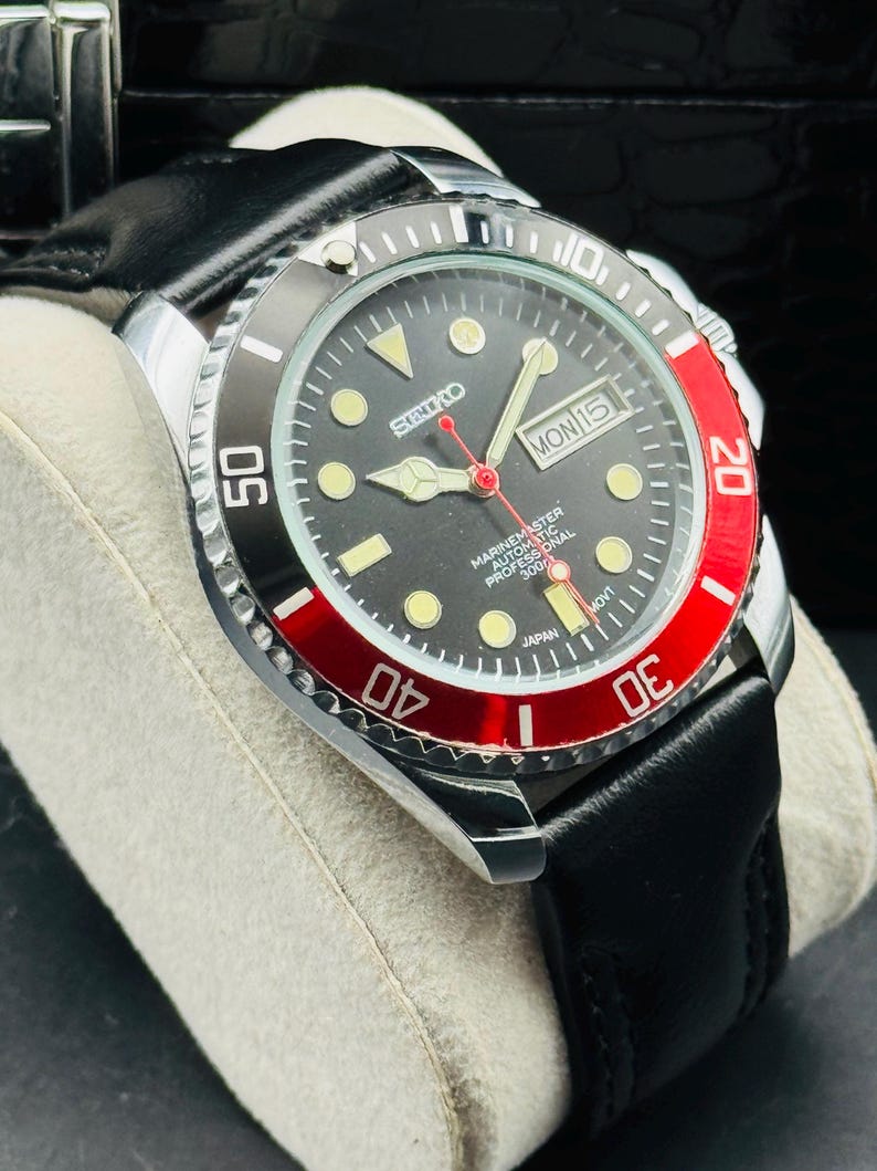 May include: A black and red Seiko Marinemaster automatic watch with a black leather strap. The watch face is black with luminous markers and hands. The bezel is black and red, with white numerals. The day and date are displayed at the 3 o'clock position.