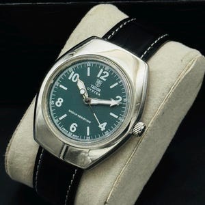 May include: A vintage Tudor Oyster watch with a silver case and a dark green dial. The watch has white hour markers and hands, a black leather strap with white stitching, and a crown. The watch is displayed on a cream-coloured cushion.