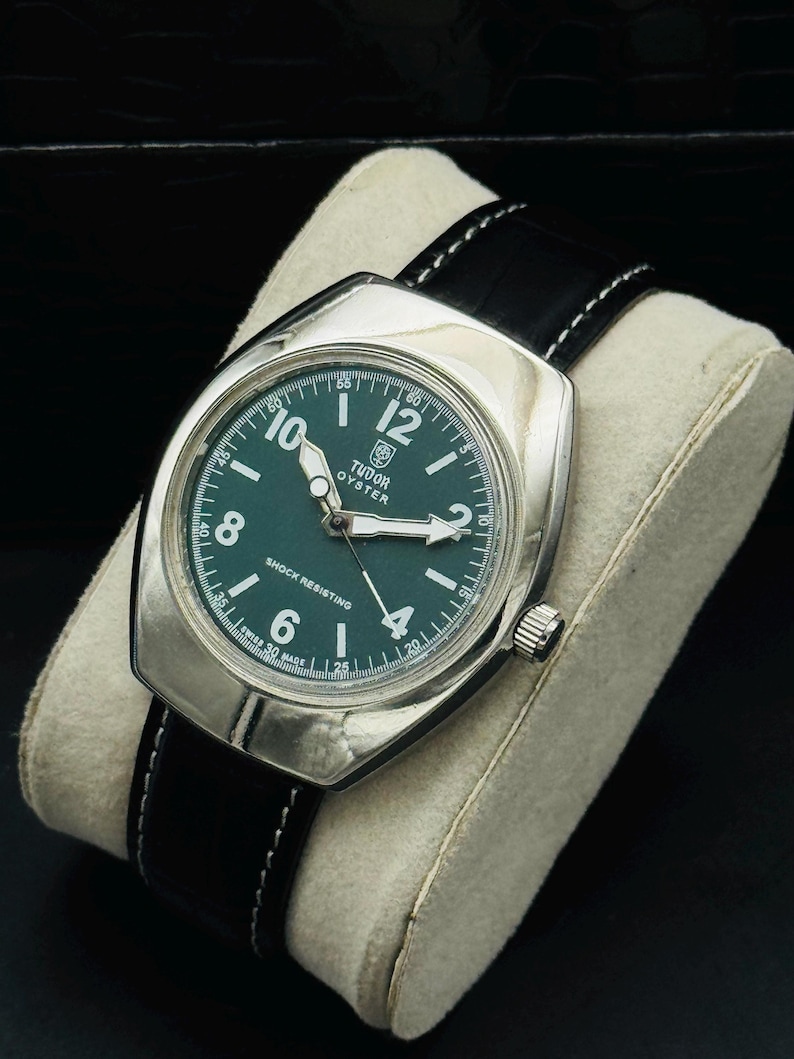 May include: A vintage Tudor Oyster watch with a silver case and a dark green dial. The watch has white hour markers and hands, a black leather strap with white stitching, and a crown. The watch is displayed on a cream-coloured cushion.