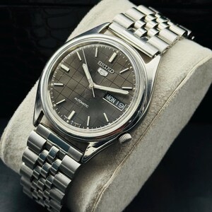 Op de afbeelding: Een zilverkleurig Seiko 5 automatisch horloge met een grijze geruite wijzerplaat. Het horloge heeft een datumweergave en een roestvrijstalen armband. Het horloge wordt gepresenteerd op een grijs kussen.