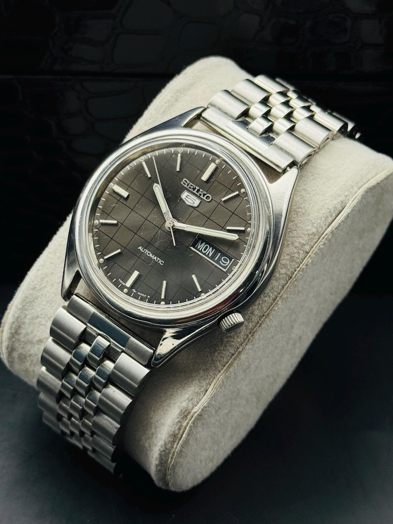 Op de afbeelding: Een zilverkleurig Seiko 5 automatisch horloge met een grijze geruite wijzerplaat. Het horloge heeft een datumweergave en een roestvrijstalen armband. Het horloge wordt gepresenteerd op een grijs kussen.