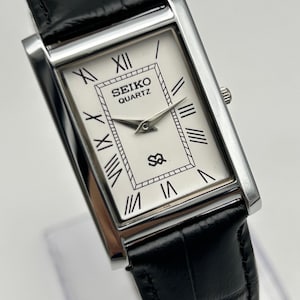 Puede incluir: Reloj Seiko Quartz rectangular con esfera blanca, n&uacute;meros romanos negros y correa de cuero negro. El reloj tiene agujas plateadas y una caja plateada. La correa es de cuero negro.