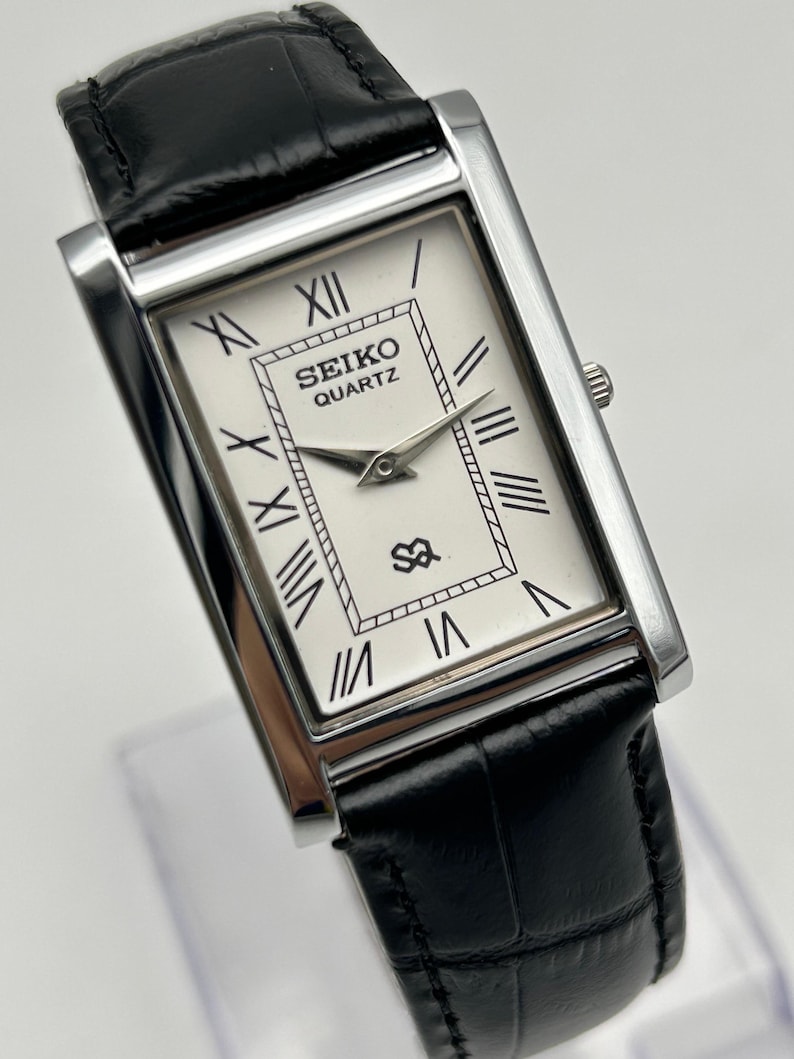 Puede incluir: Reloj Seiko Quartz rectangular con esfera blanca, n&uacute;meros romanos negros y correa de cuero negro. El reloj tiene agujas plateadas y una caja plateada. La correa es de cuero negro.
