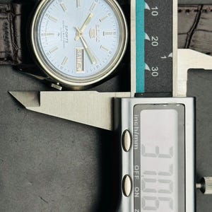 May include: A silver-toned wristwatch with a light blue face and a brown leather strap. The watch face displays the day and the word "SPORTS". The digital caliper shows a measurement of 37.06 mm.