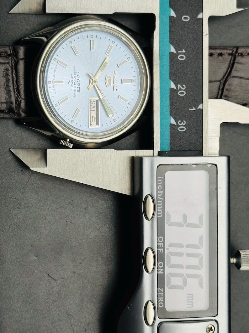 May include: A silver-toned wristwatch with a light blue face and a brown leather strap. The watch face displays the day and the word "SPORTS". The digital caliper shows a measurement of 37.06 mm.