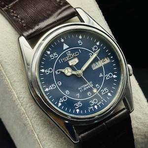 May include: A vintage Seiko 5 automatic watch with a dark blue dial and silver-tone case. The watch features a day and date display, luminous hands, and a brown leather strap. The watch face displays the text "SEIKO 5 AUTOMATIC 21 JEWELS".