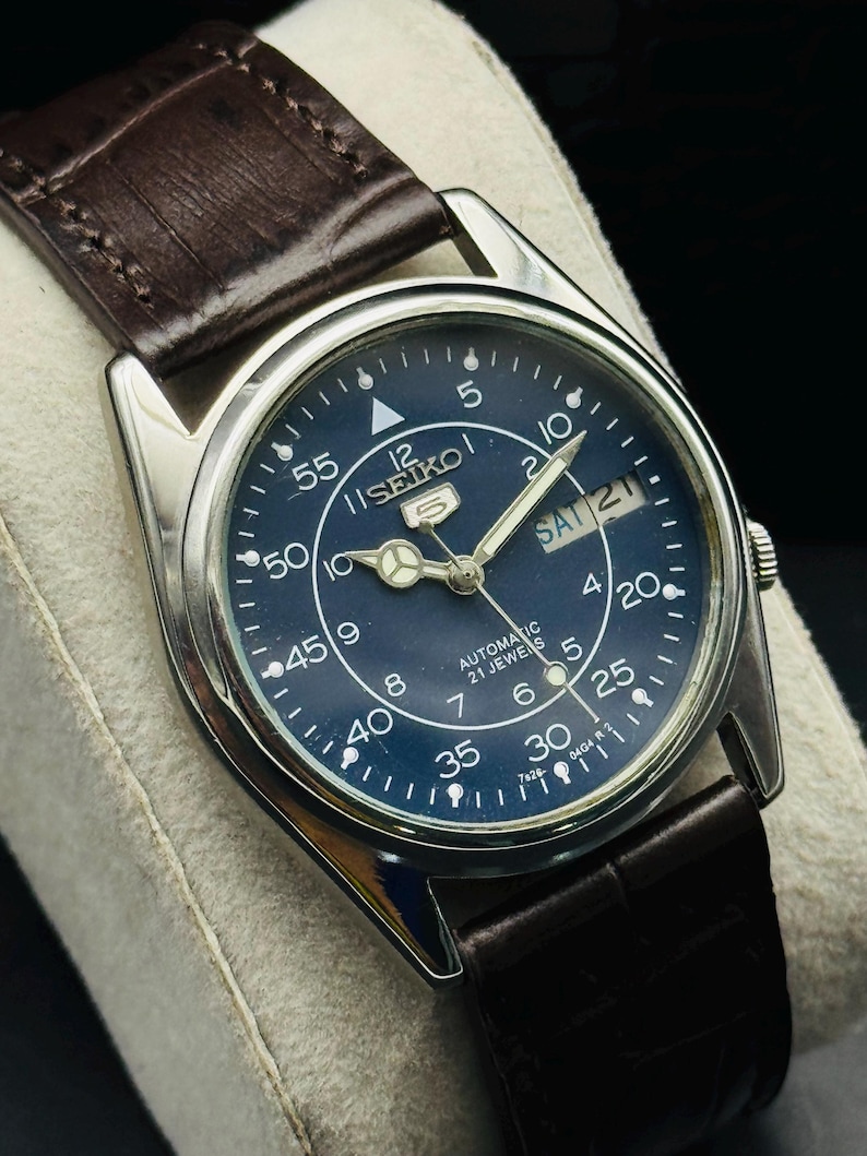 May include: A vintage Seiko 5 automatic watch with a dark blue dial and silver-tone case. The watch features a day and date display, luminous hands, and a brown leather strap. The watch face displays the text "SEIKO 5 AUTOMATIC 21 JEWELS".