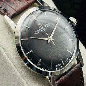 May include: A silver-toned Jaeger-LeCoultre Club watch with a dark gray face and silver hands and markers. The watch has a brown leather band. The watch face reads "Jaeger-LeCoultre Club" and "Swiss Made".