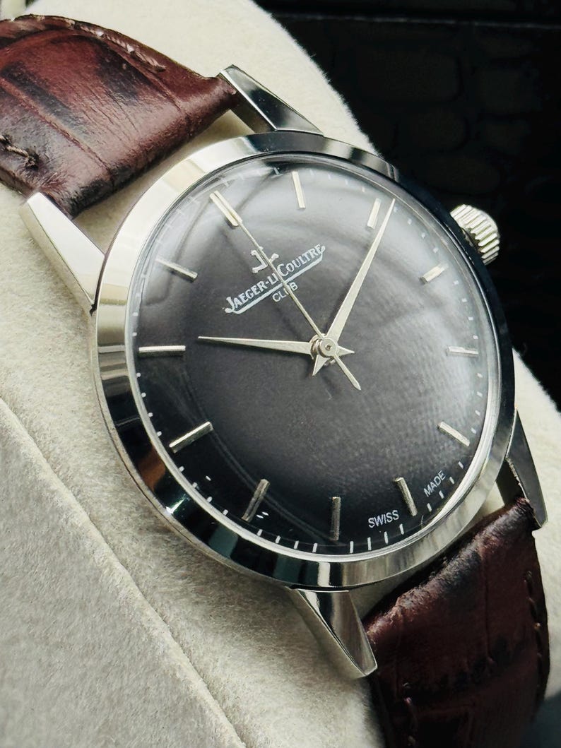 May include: A silver-toned Jaeger-LeCoultre Club watch with a dark gray face and silver hands and markers. The watch has a brown leather band. The watch face reads "Jaeger-LeCoultre Club" and "Swiss Made".