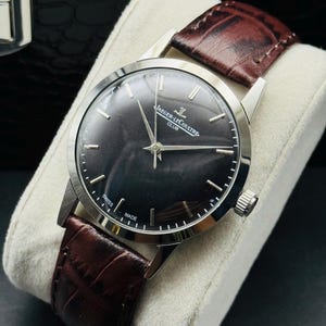 May include: A silver-toned Jaeger-LeCoultre Club watch with a dark gray face and a brown leather band. The watch has silver hands and markers, and the band is textured. The watch is displayed on a white cushion.