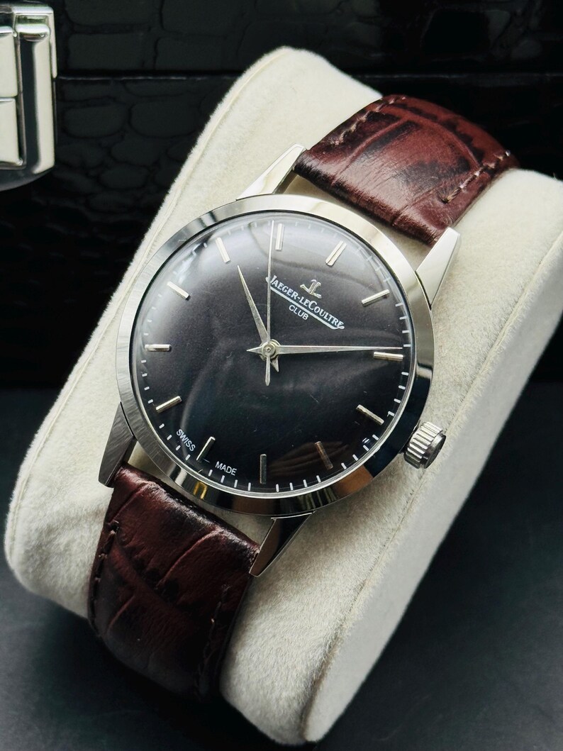 May include: A silver-toned Jaeger-LeCoultre Club watch with a dark gray face and a brown leather band. The watch has silver hands and markers, and the band is textured. The watch is displayed on a white cushion.