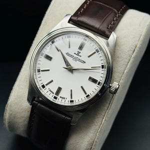 May include: A silver-toned wristwatch with a white face and a brown leather band. The watch features the text "JAEGER-LECOULTRE CLUB" and "SWISS MADE". The watch is displayed on a light-colored cushion.