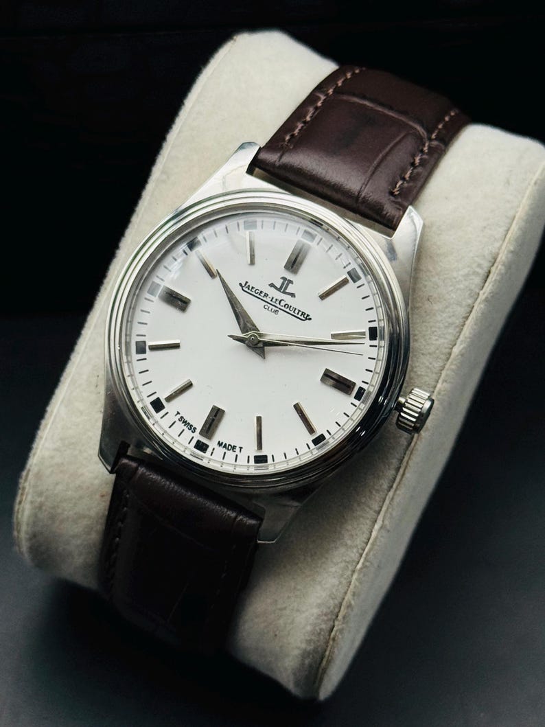 May include: A silver-toned wristwatch with a white face and a brown leather band. The watch features the text "JAEGER-LECOULTRE CLUB" and "SWISS MADE". The watch is displayed on a light-colored cushion.