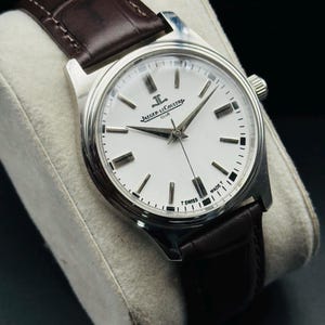 May include: A silver-toned wristwatch with a white face and black markers. The watch features a brown leather band and the text "JAEGER-LECOULTRE CLUB" on the face. The watch is displayed on a gray cushion.