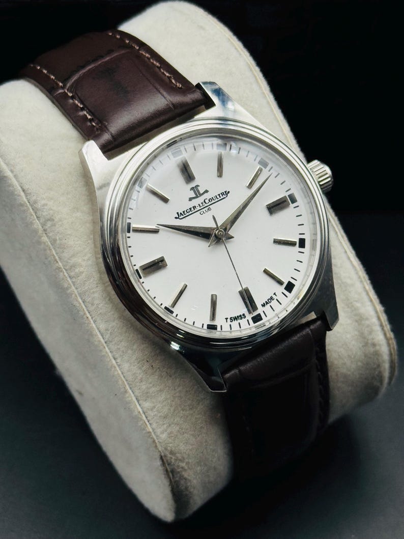 May include: A silver-toned wristwatch with a white face and black markers. The watch features a brown leather band and the text "JAEGER-LECOULTRE CLUB" on the face. The watch is displayed on a gray cushion.