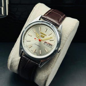 May include: A silver-toned Seiko automatic watch with a brown leather strap. The watch face is silver with gold hands and markers, and a red second hand. The date is displayed at the 3 o'clock position. The watch is displayed on a cream-colored cushion.