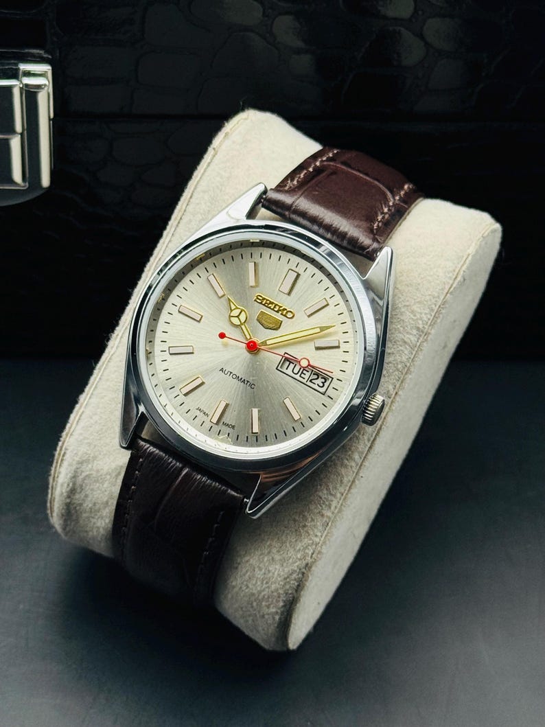 May include: A silver-toned Seiko automatic watch with a brown leather strap. The watch face is silver with gold hands and markers, and a red second hand. The date is displayed at the 3 o'clock position. The watch is displayed on a cream-colored cushion.