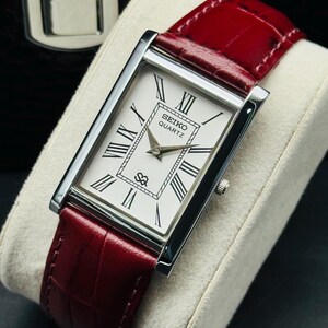 May include: A rectangular Seiko Quartz watch with a white face, black Roman numerals, and a red leather band. The watch has a silver-colored case and a small seconds dial. The watch is displayed on a cream-colored cushion.