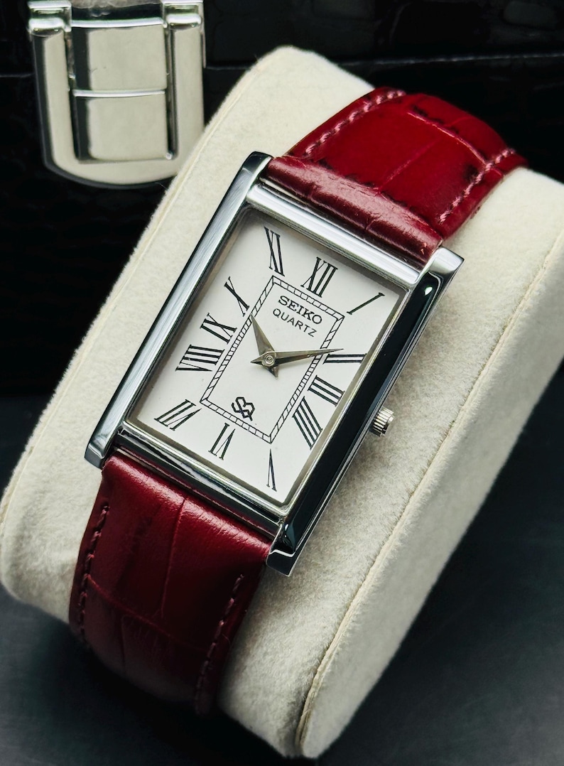 May include: A rectangular Seiko Quartz watch with a white face, black Roman numerals, and a red leather band. The watch has a silver-colored case and a small seconds dial. The watch is displayed on a cream-colored cushion.