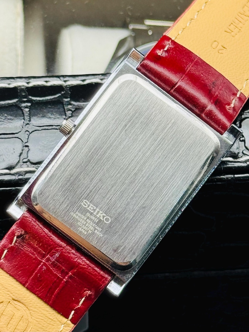 May include: A rectangular Seiko quartz watch with a silver-toned back and a red leather strap. The watch has a silver crown and the back is engraved with "SEIKO QUARTZ WATER RESISTANT STAINLESS STEEL BACK 7N43-0070 JAPAN".